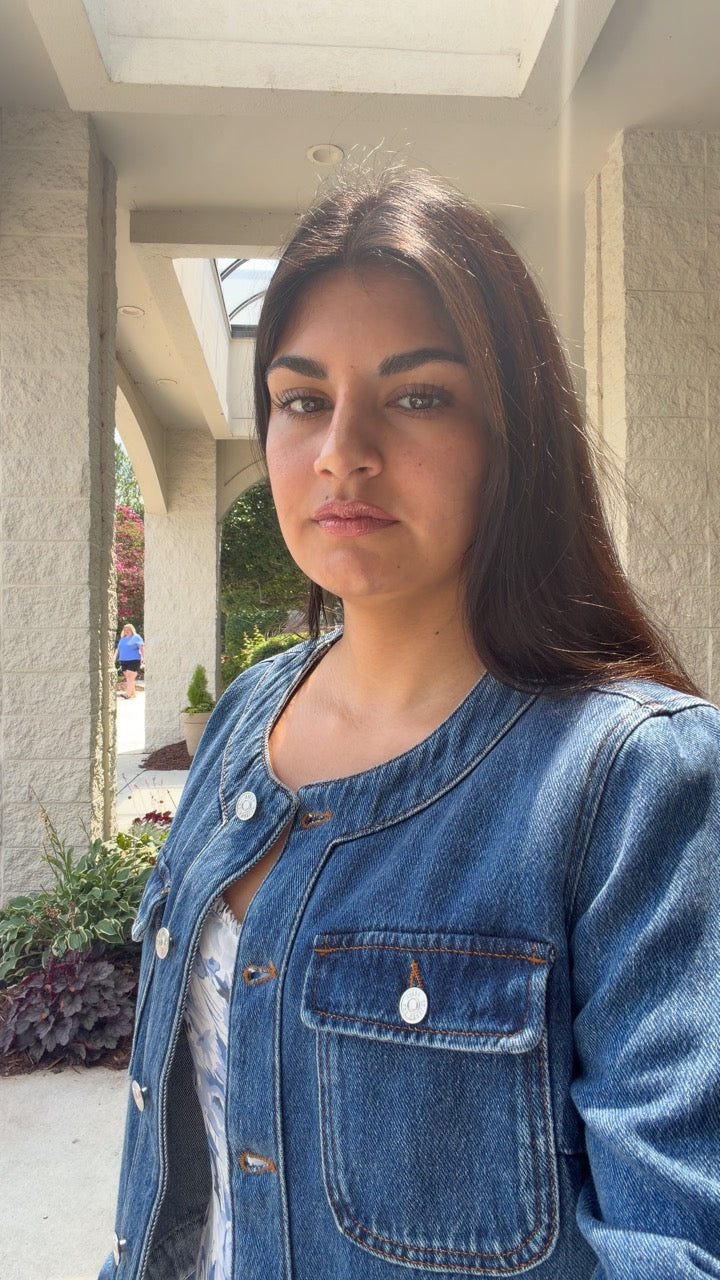 A woman wearing a blue denim jacket over a white top, standing in a covered outdoor walkway.