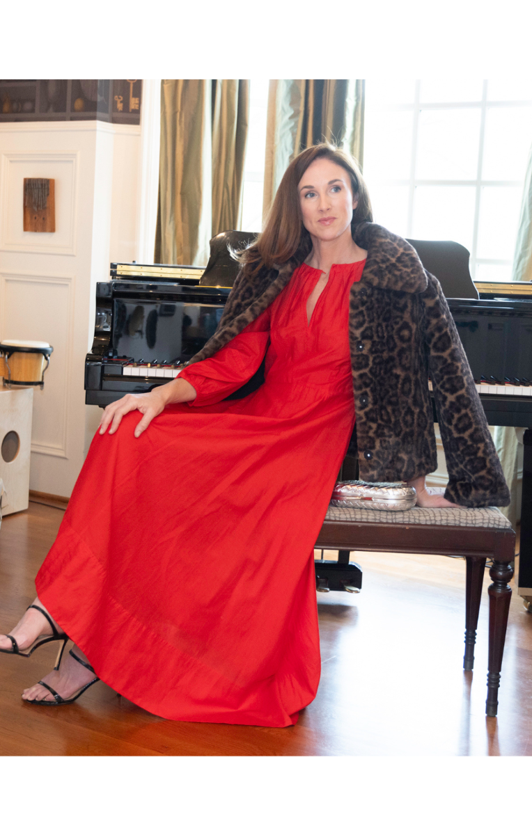 Woman in a red dress and leopard print coat sitting in front of a piano.